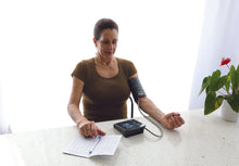 Load image into Gallery viewer, Patient sitting at a table and taking her blood pressure with the BD252 monitor